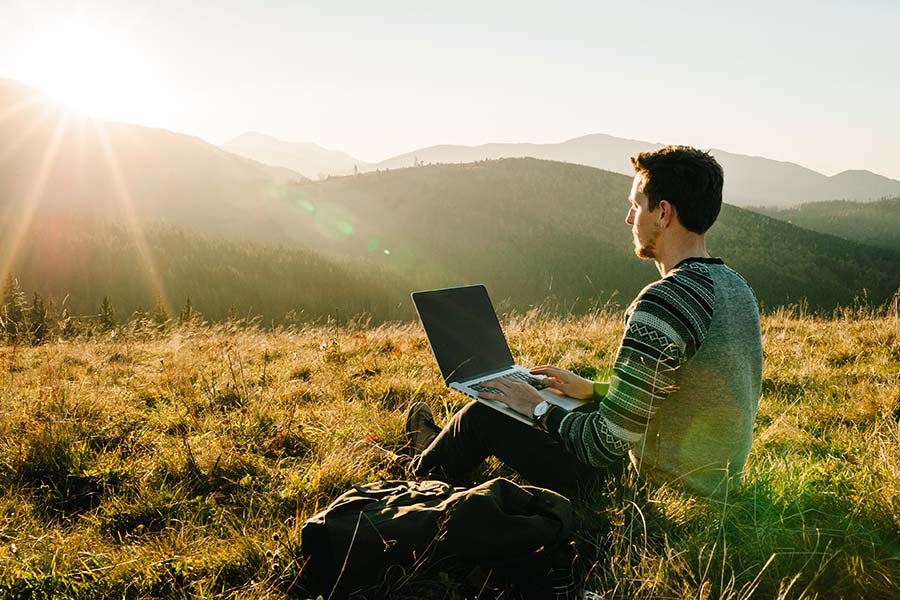 Blog - Portrait of a Young Man Sitting on the Grass Using a Laptop While on a Hike in the Mountains at Sunset
