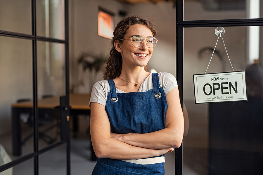 Business Insurance - Closeup Portrait of a Cheerful Young Business Owner Standing Outside Her Store Next to an Open Sign